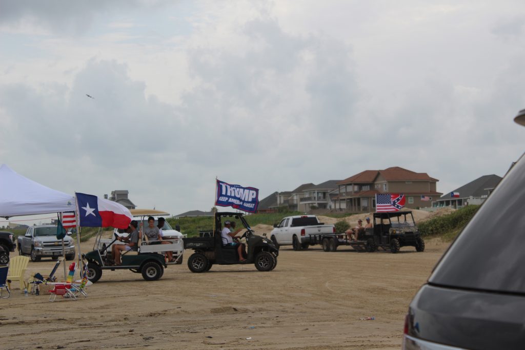 A beach with trucks and ATVs. An American flag, Confederate flag hybrid is seen on one ATV. A "Trump. Keep America Great" flag on another.