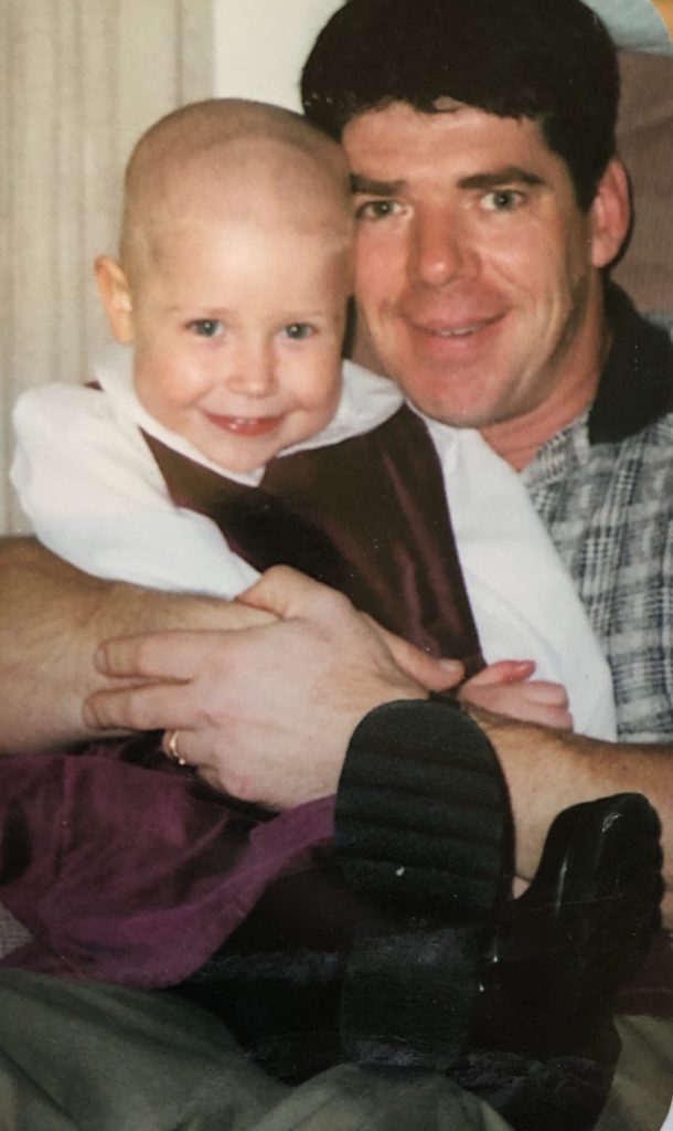 Sarah with no hair sitting on her dad's lap as they both smile at the camera.