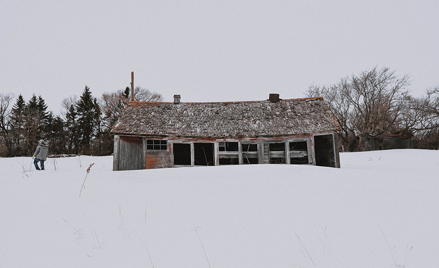 A man stands beside a dilapidated house in a snowy field.