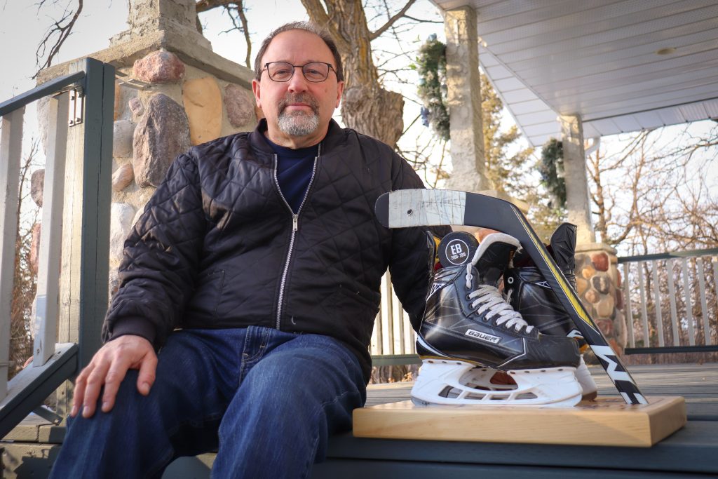 A middle aged man sits on the steps of a porch beside a pair of skates and the blade of a hockey stick that have been mounted on a piece of wood.