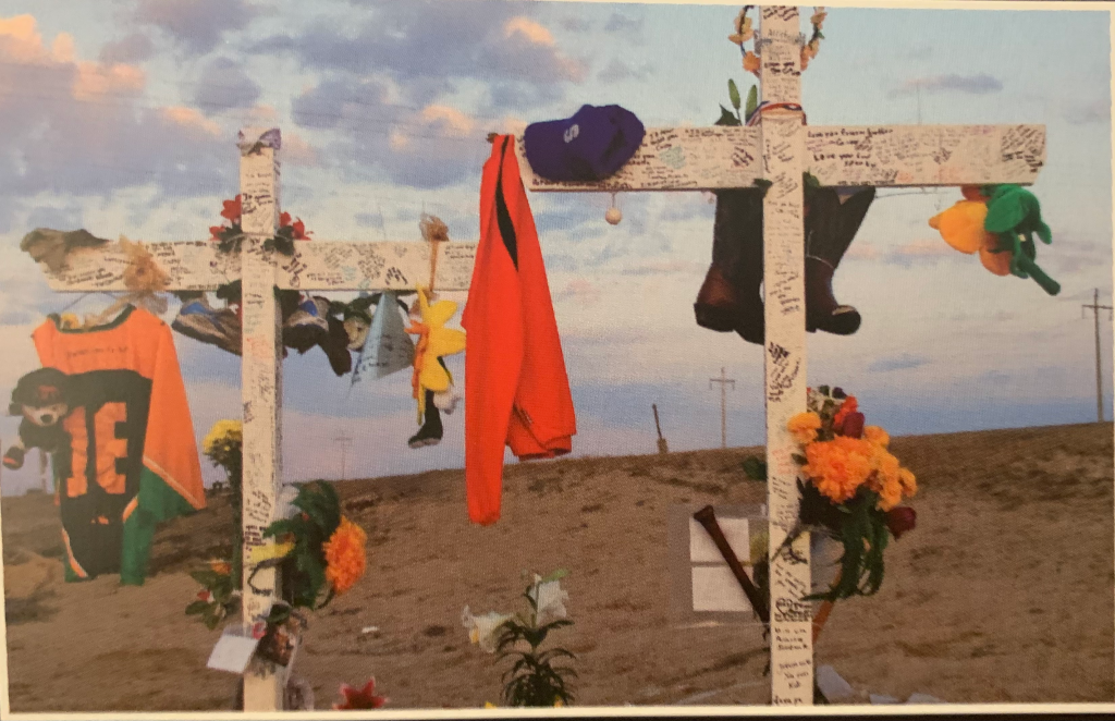 Two white crosses by the roadside covered in writing, flowers, orange jerseys and more.