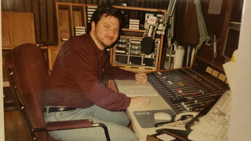 A on older film photo of a man sitting at a microphone and analog sound board.