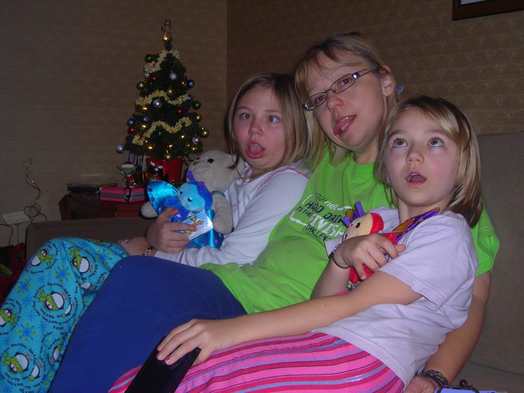 A woman with her arms around two young girls all sit on a couch. They all wear pajamas. The room is decorated for Christmas.