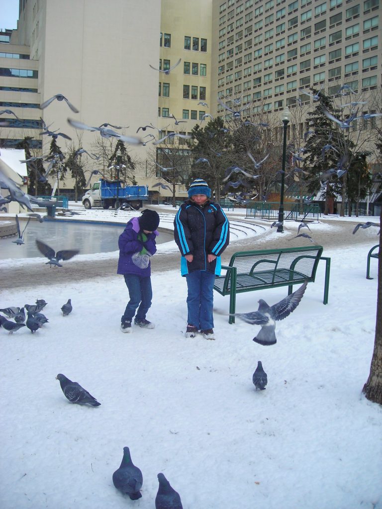 Two young girls in winter wear are surrounded by a flock of pigeons as they stand in a snowy city park.