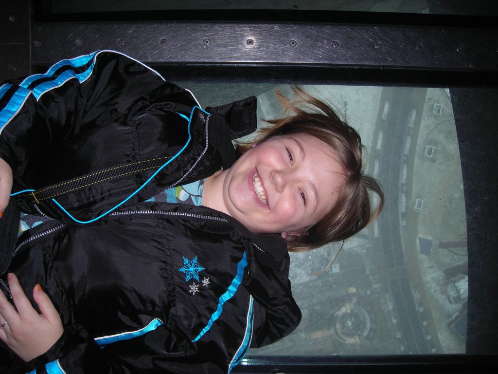 Sydney smiling at the camera as she lays down on a glass floor, with city seen below.