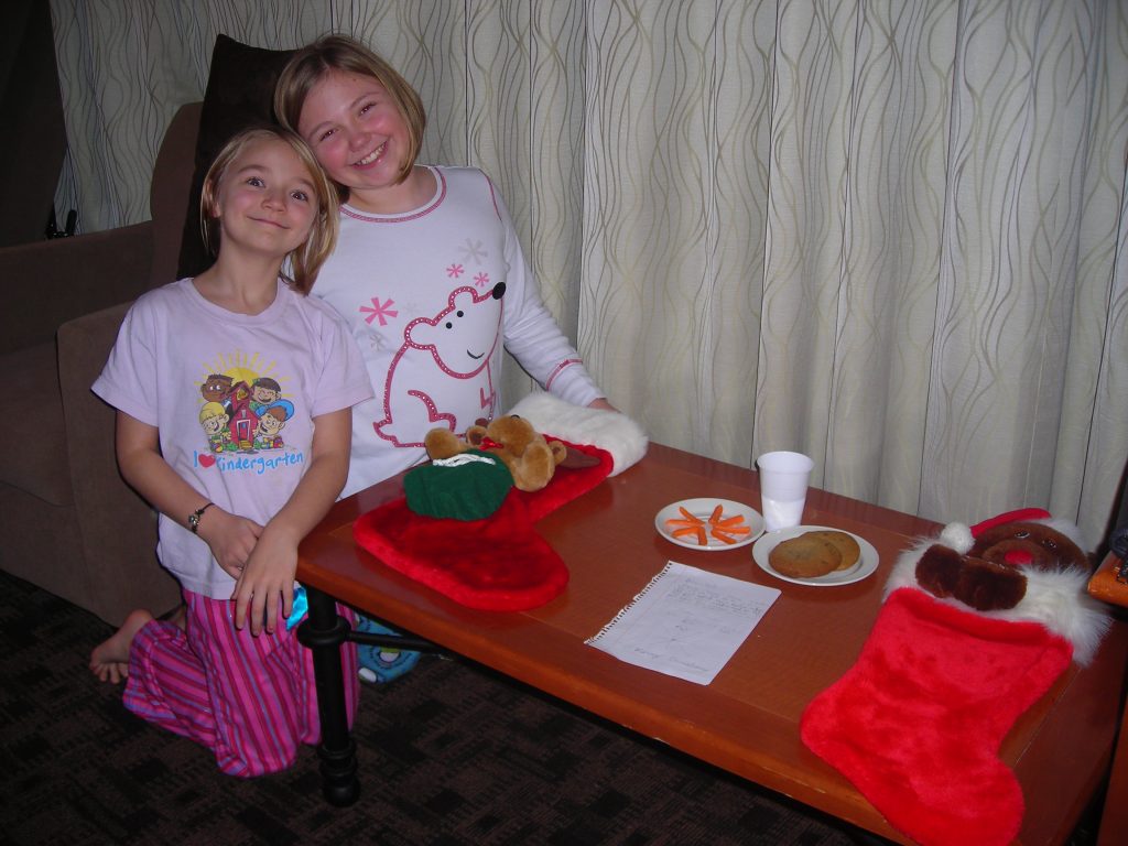 Two young girls in pajamas pose beside a table with Christmas stockings, milk, cookies, carrots and a note.