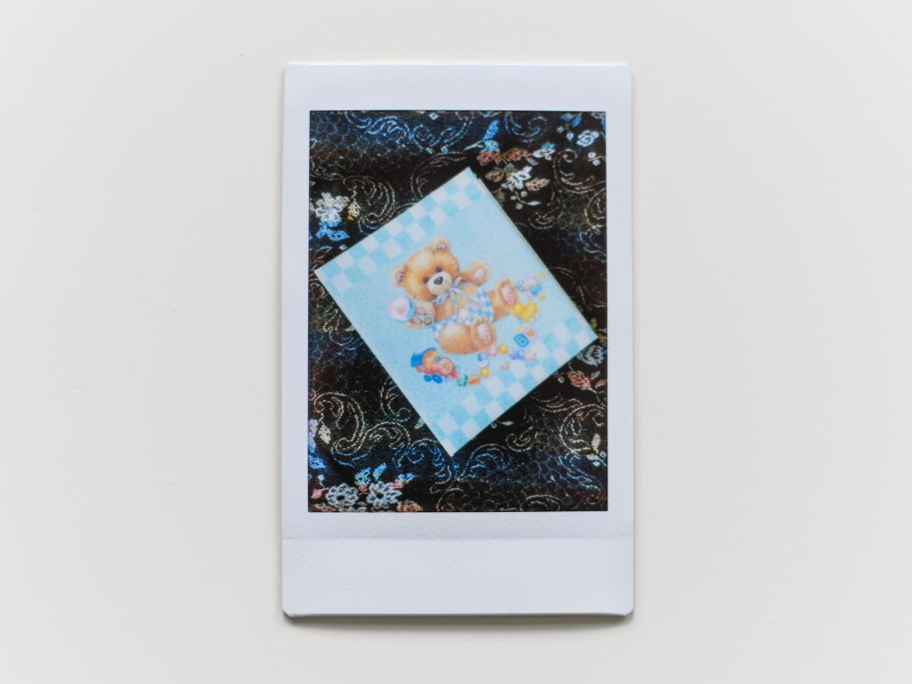 A polaroid of a book with a teddy bear dressed like a baby on it.