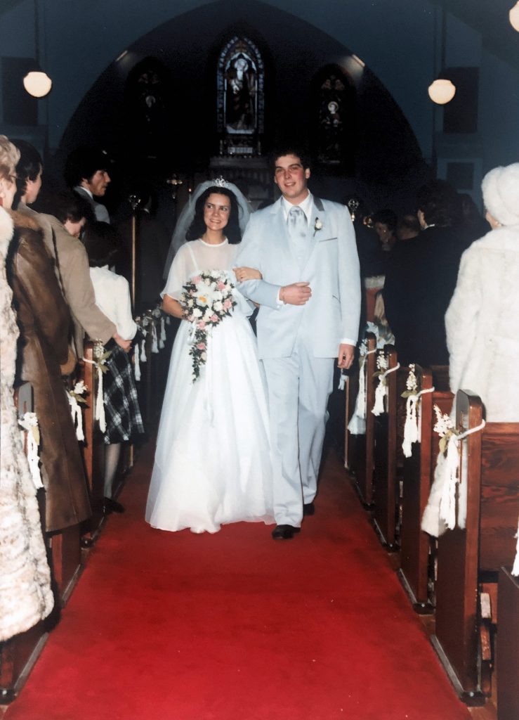 Pam and Bruce walking down the aisle of a church dressed in a wedding dress and baby blue tuxedo.
