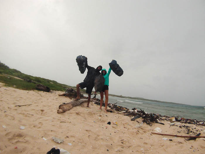 A man and a woman in raincoats on a beach hold up full garbage bags.