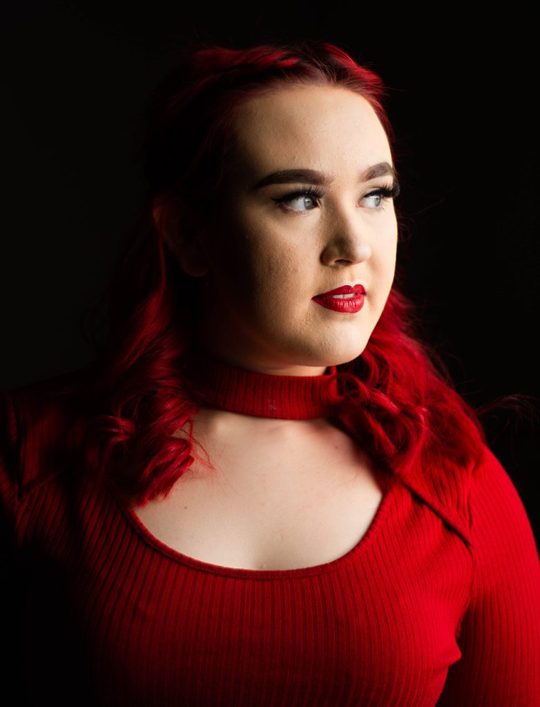 A portrait of Harley in dramatic lighting. Her hair, top, and lips are all a bold red.