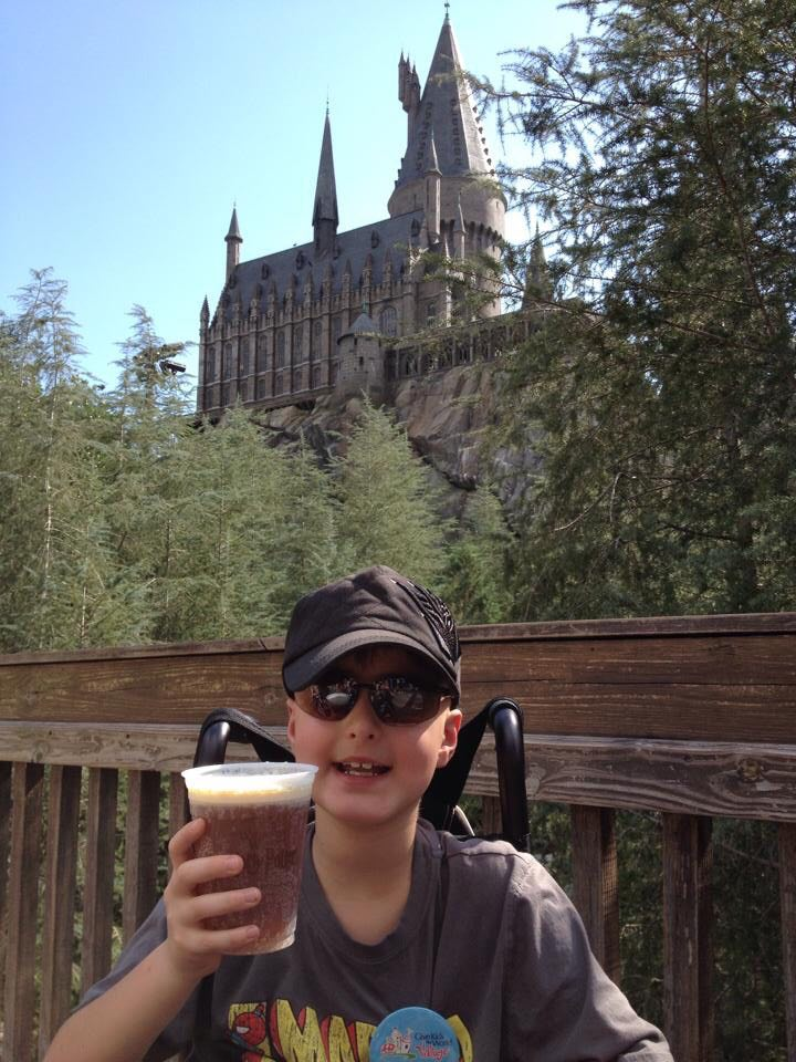 Madox in front of Hogwarts Castle at The Wizarding World of Harry Potter in Orlando. He is holding a cup of Butterbeer and is sitting in a wheelchair. 