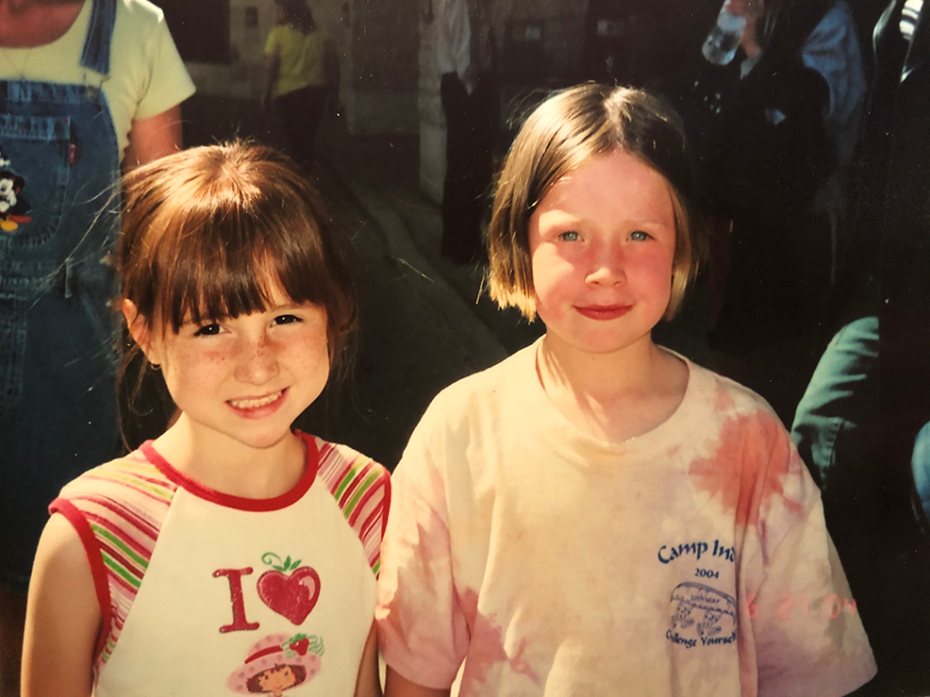 Sarah and her friend Caitlyn as young children smiling at the camera.