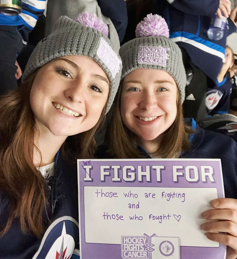 Sarah and her friend Caitlyn at a Jet's hockey game wearing matching "hockey fights cancer" toques and Jet's jerseys. Sarah holds a sign that reads "I fight for those who are fighting and those who fought".