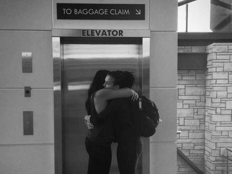 Sam and Taylor Toews hug each other in the airport in front of the elevator and baggage claim sign.
