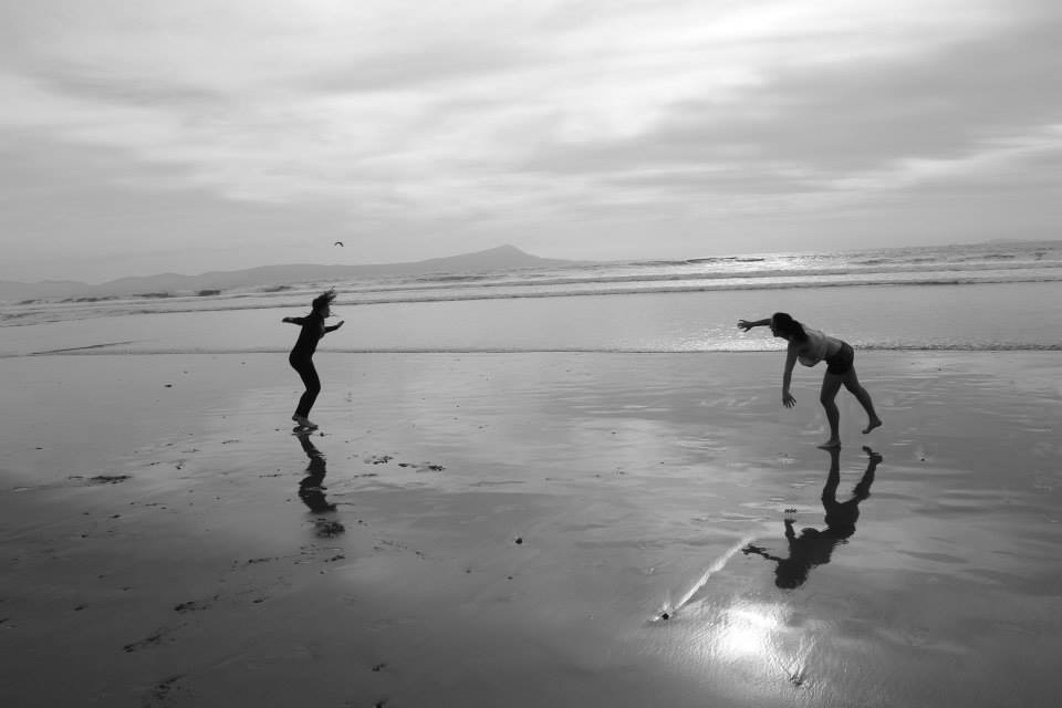 Two women dance on a beach with the waves creeping in on them gently.
