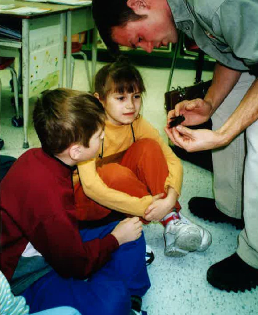 Liliya sitting on the floor with her cousin as children looking at a spider a man is holding.