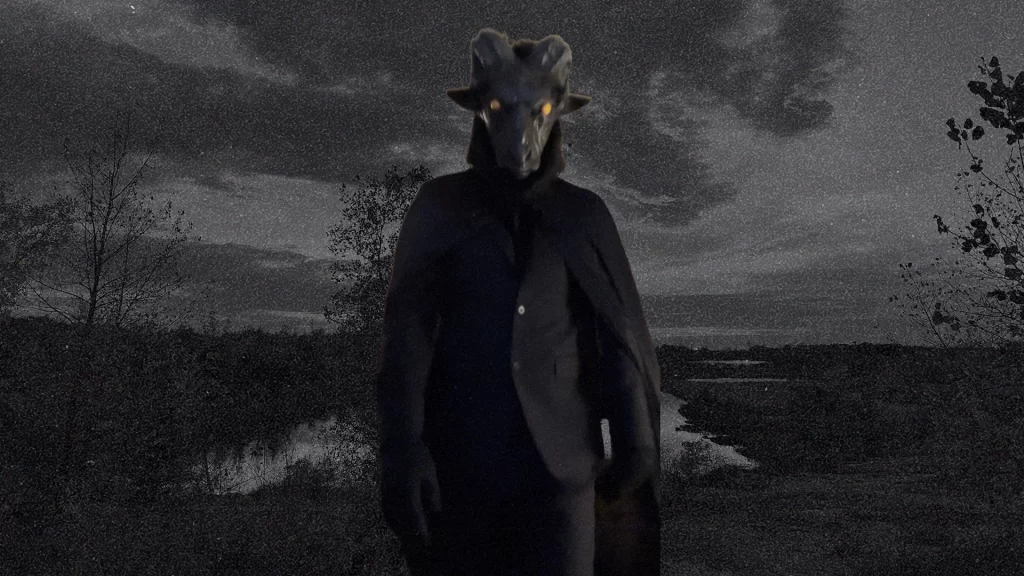 A goat-headed figure with glowing eyes in a black suit stands in the foreground. In the background, a grainy landscape with a small body of water.