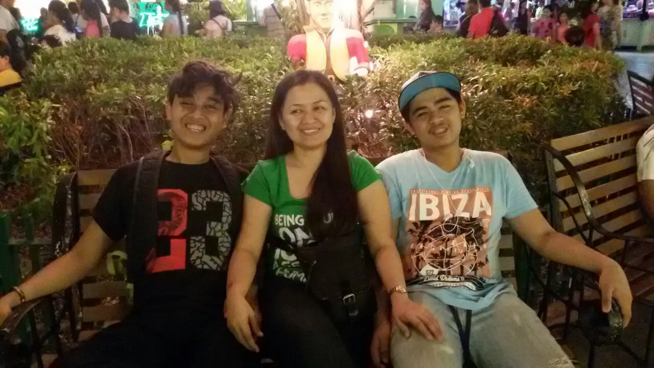 Mom and her two sons sat on a bench with shrubs and people in the background.