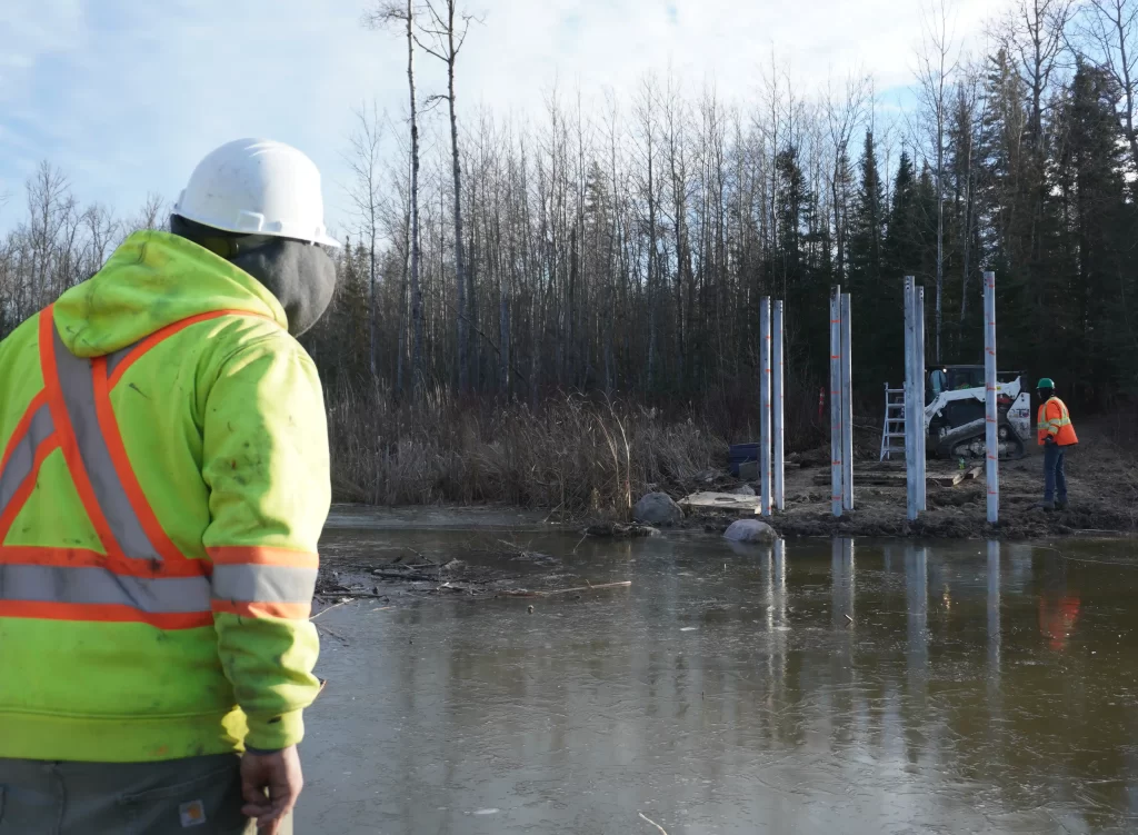 Morley Daniels stands looking over the pond at the Otter Falls jobsite. He's wearing a bright yellow and orange high-vis jacket.