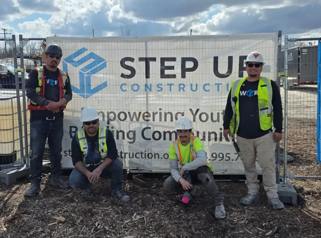Four men stand in front of a construction site, and they're all wearing high vis vests and hard hats. A sign behind them reads "Step Up Construction" beside the Step Up Construction logo.