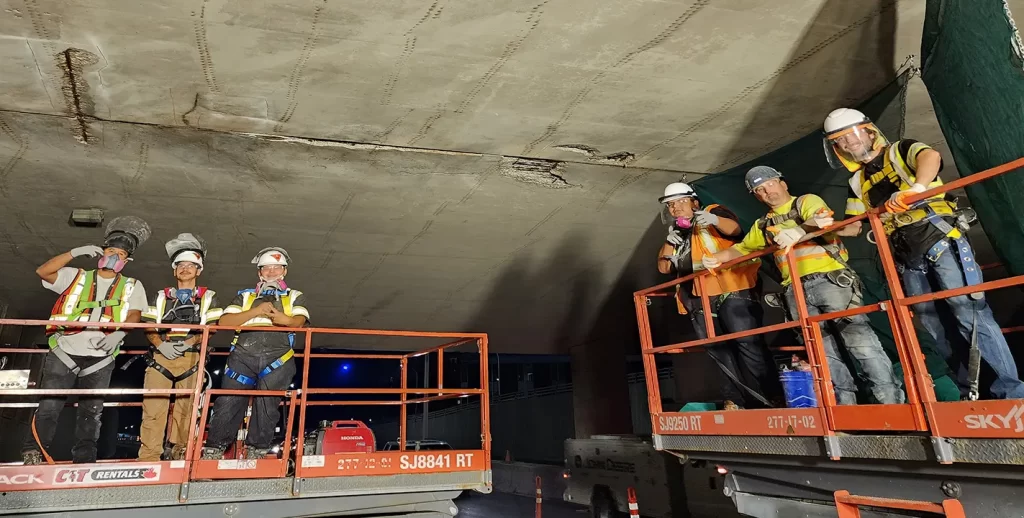 A group of six crew members working the night shift pose for a photo on lifts underneath the bridge at the St. James jobsite.