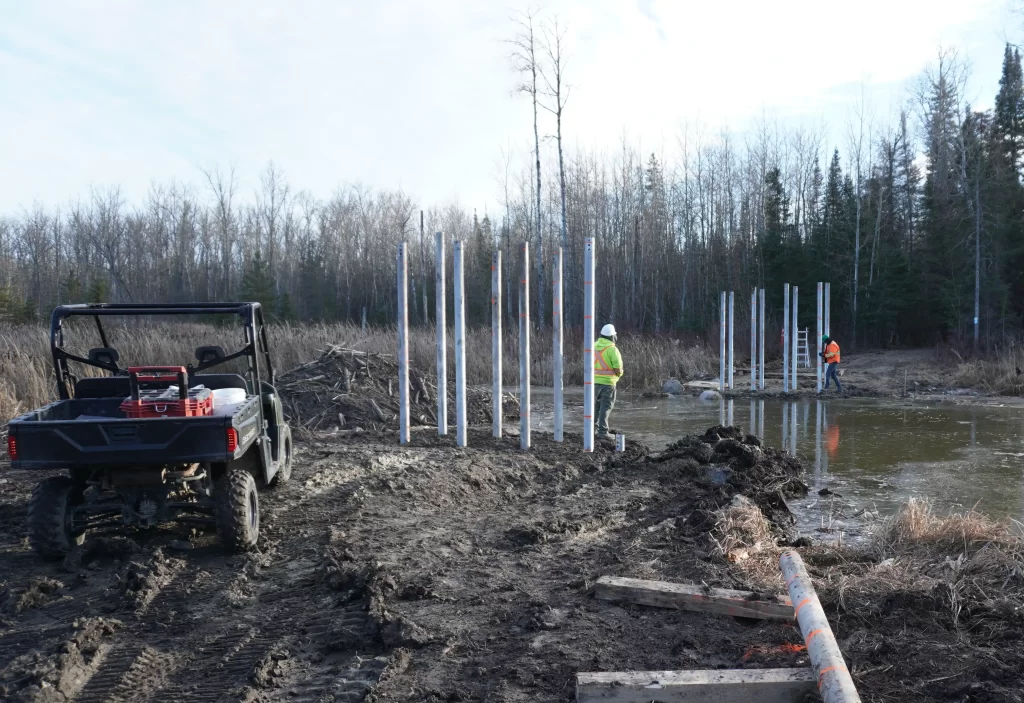 A zoomed-out view shows Morley Daniels wearing a high-vis jacket at the Otter Falls jobsite. Several metal posts stick out of the ground, forming the foundation for the bridge being built.