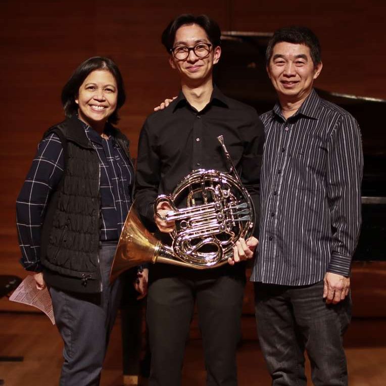 Three people standing on stage, the middle person holds a French Horn.