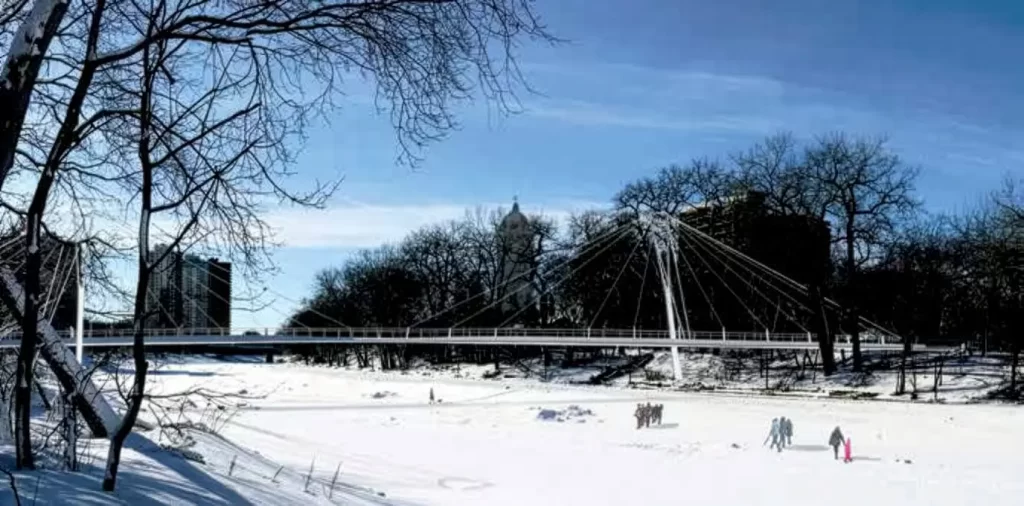 A digitally-rendered suspension footbridge crosses a frozen Assiniboine River with pedestrians on the ice.