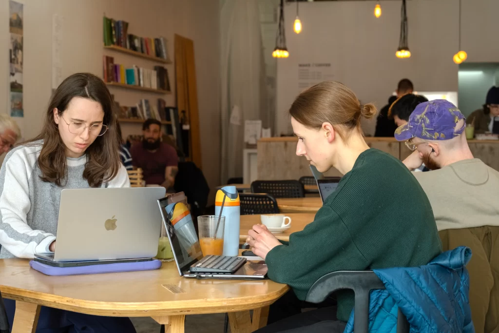 Two young women sit at a triangular table made of plywood in a cafe working at their laptops. More patrons are sitting in the background with hanging light fixtures overhead.