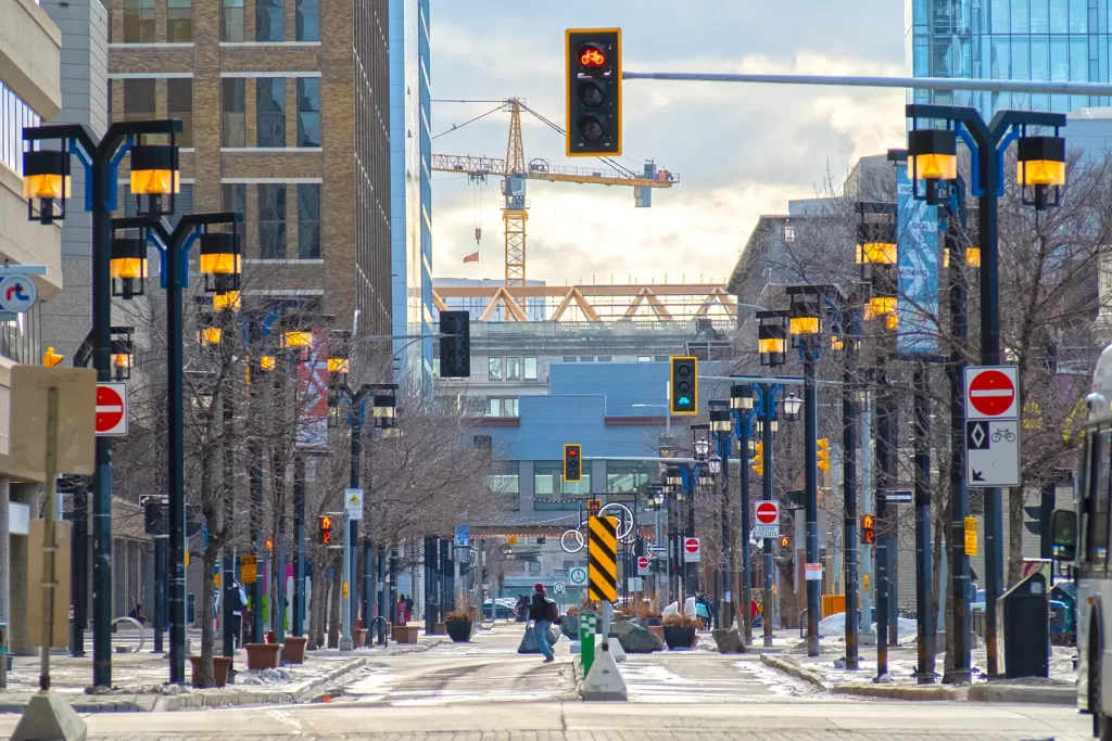 There are a few pedestrians on a slightly snowy Graham Avenue. Trees, lamp posts, and large buildings line both sides and a crane stands at the end.