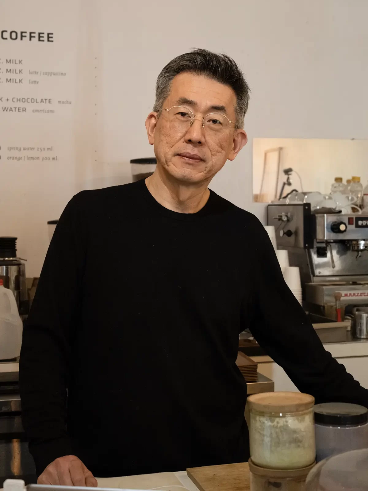 A middle-aged Korean man stands behind a bar with a jar of matcha in front of him and an espresso machine behind him.
