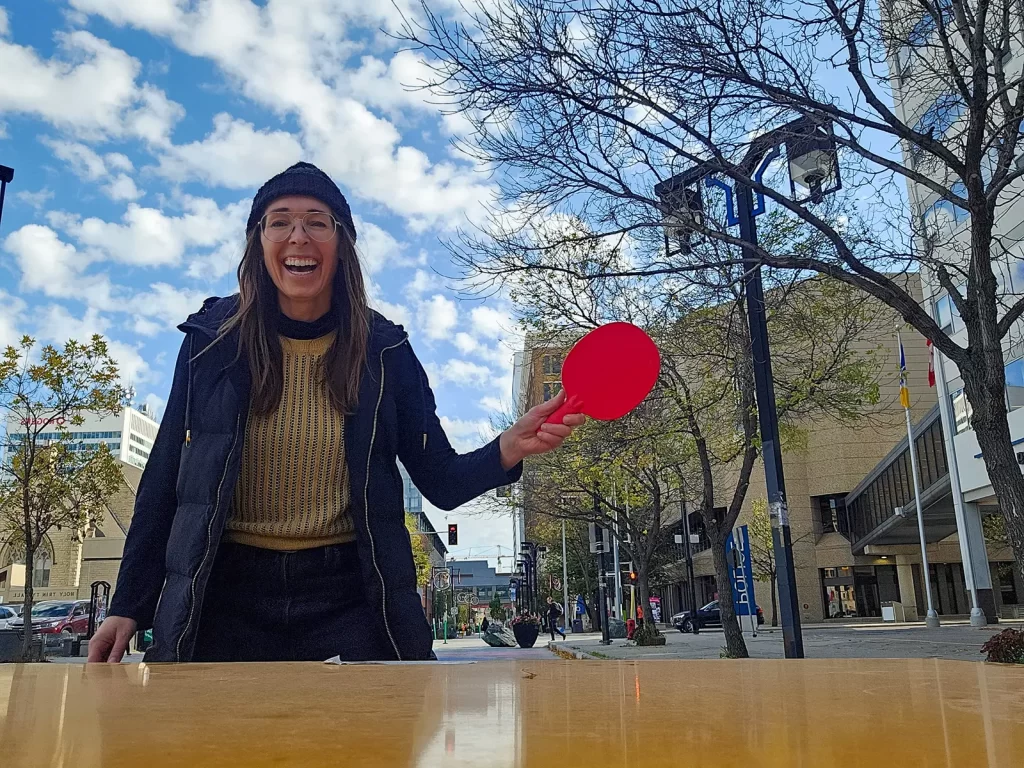 A woman smiles holding a bright red table tennis paddle wearing a jacket and tuque on a downtown street under a partly cloudy sky.