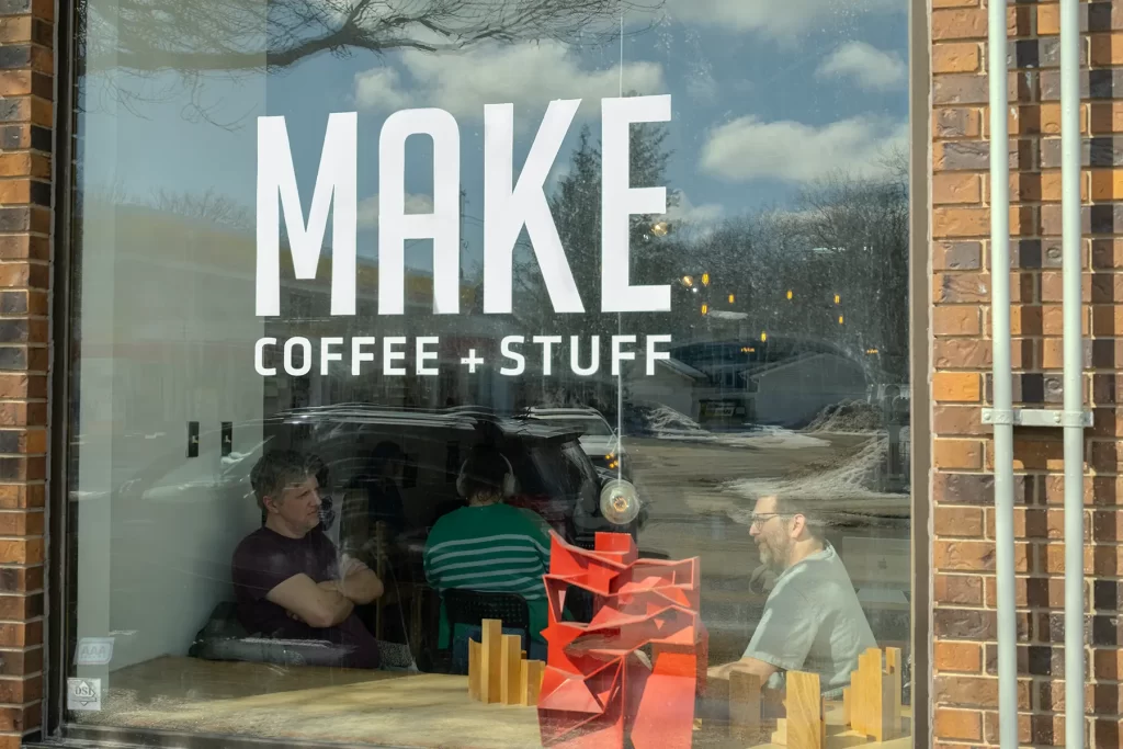 Two men sit across the table from each other seen through the front window of MAKE Coffee + Stuff. Design mockups are displayed in the window.