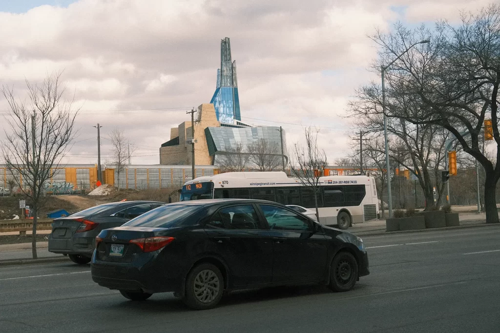 Cars, a bus, and a train pass each other at Main Street with the Canadian Museum for Human Rights towering in the background.