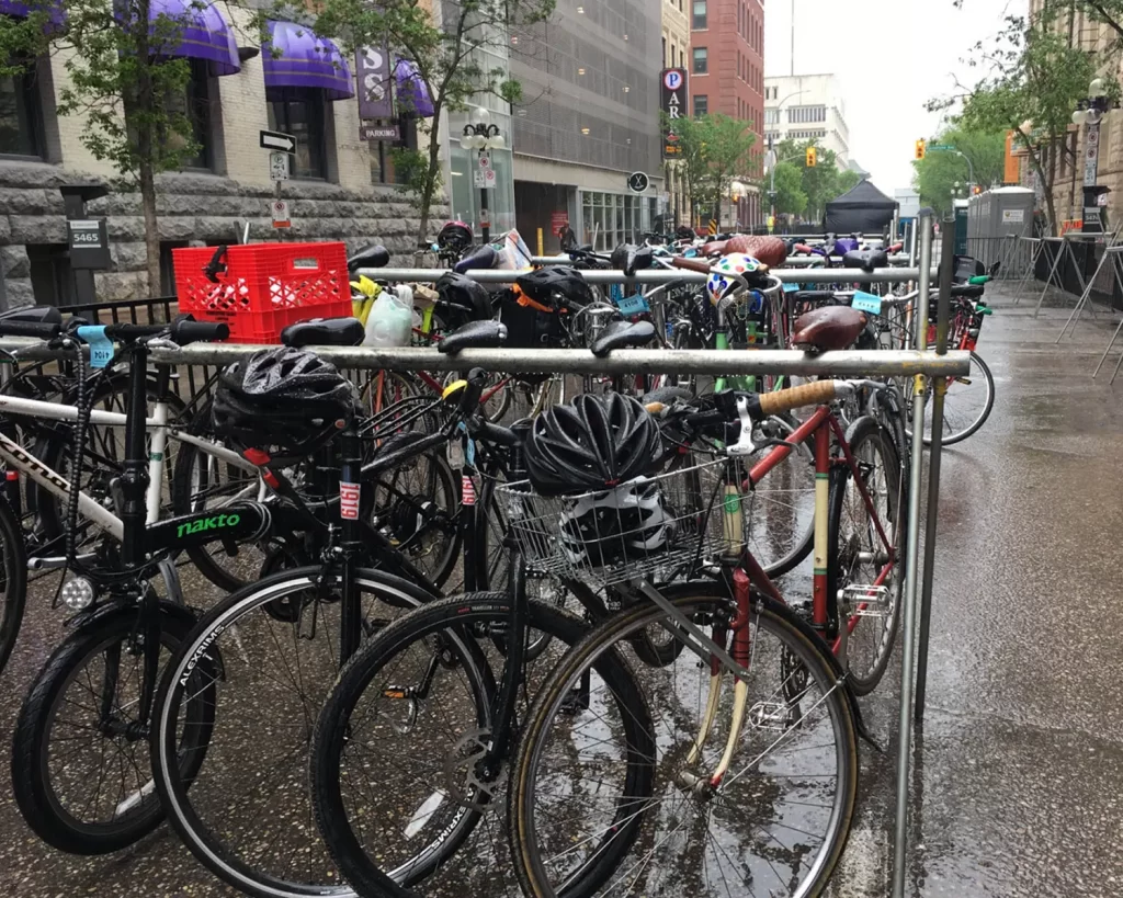 Six bikes hang by their saddles from a steel rack on a wet street. Several more full bike racks are lined up in the background and several empty racks are lined up to the side.